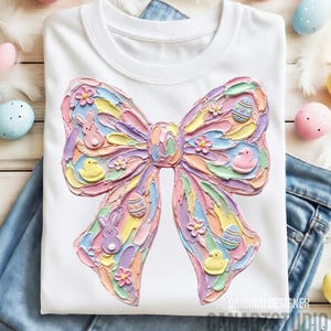 May include: White t-shirt featuring a large, textured bow design in pastel colors. The bow is adorned with Easter-themed embellishments, including eggs, bunnies, and ducklings. The shirt is displayed on a pair of blue jeans.