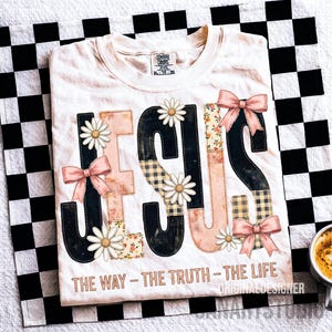 May include: Cream-colored t-shirt with the word "JESUS" in a patchwork design. The letters are black, pink, and floral, with daisy accents and pink bows. The text "THE WAY - THE TRUTH - THE LIFE" is printed below.