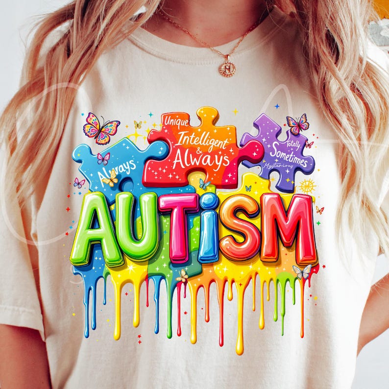 May include: A white t-shirt with a colorful graphic design of the word "AUTISM" in dripping paint. The design includes puzzle pieces with the text "Unique, Intelligent & Always, Totally Sometimes Mysterious".