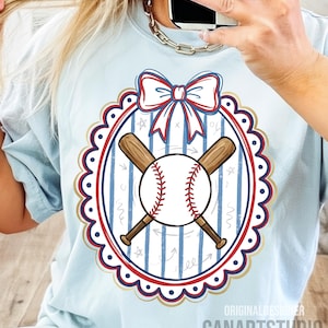 May include: Light blue t-shirt featuring a baseball design. The design includes a baseball, crossed bats, and a bow, all within a scalloped oval frame. The text "CANARTSTUDIO" is visible at the bottom.