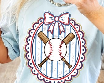 May include: Light blue t-shirt featuring a baseball design. The design includes a baseball, crossed bats, and a bow, all within a scalloped oval frame. The text "CANARTSTUDIO" is visible at the bottom.