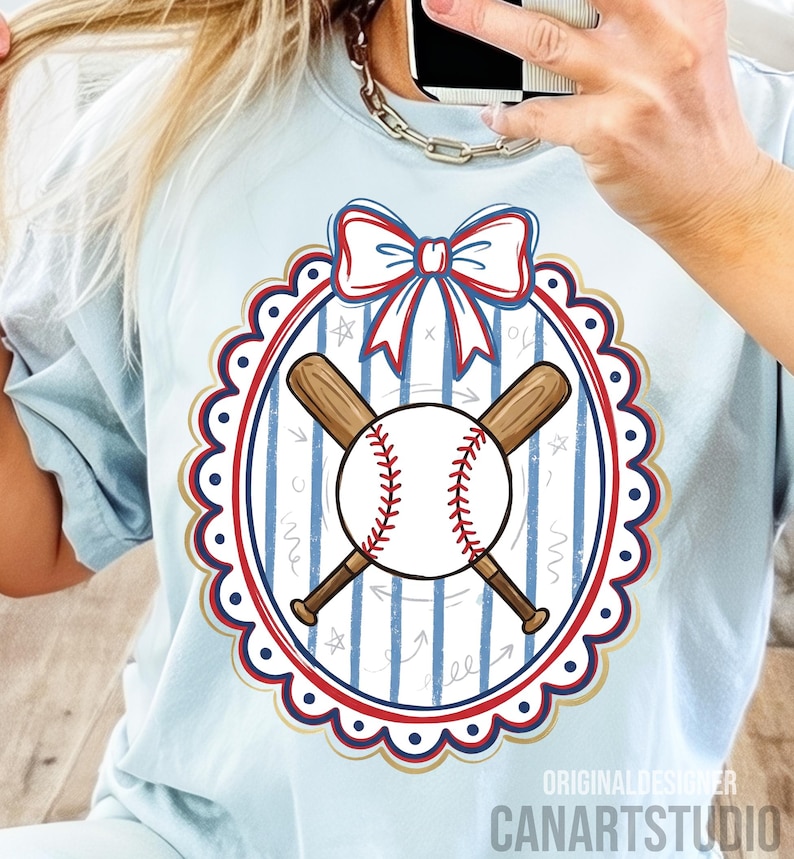 May include: Light blue t-shirt featuring a baseball design. The design includes a baseball, crossed bats, and a bow, all within a scalloped oval frame. The text "CANARTSTUDIO" is visible at the bottom.