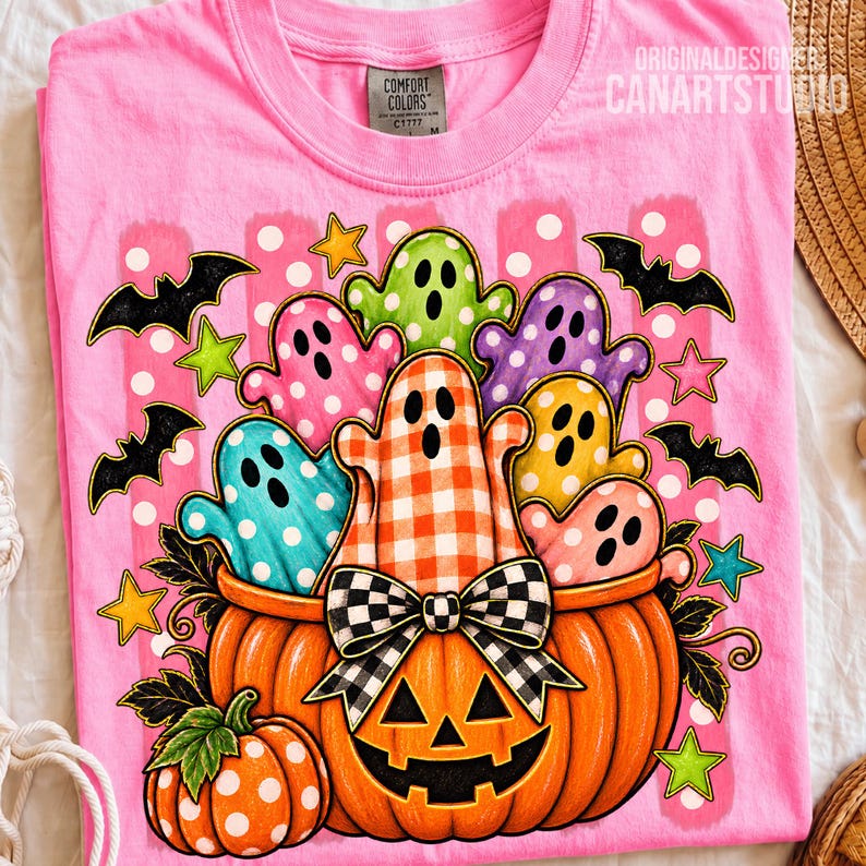 May include: A bright pink t-shirt featuring a large graphic of a carved orange pumpkin filled with colorful ghosts in various patterns. The design includes black bats, yellow and green stars, and a black and white checkered bow on the pumpkin.