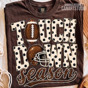 May include: A brown short-sleeved t-shirt featuring the text TOUCHDOWN SEASON in large white block letters with black polka dots. A brown football replaces the letter O, and a brown football helmet is positioned below the word TOUCH.