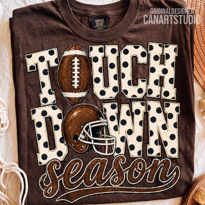 May include: A brown short-sleeved t-shirt featuring the text TOUCHDOWN SEASON in large white block letters with black polka dots. A brown football replaces the letter O, and a brown football helmet is positioned below the word TOUCH.