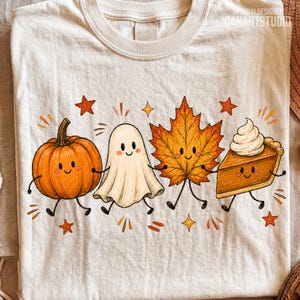 May include: A cream-colored t-shirt featuring a graphic of a smiling pumpkin, a ghost, a maple leaf, and a slice of pumpkin pie with whipped cream. The characters are holding hands, surrounded by small orange stars and dashes on the fabric.