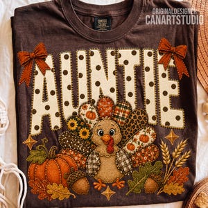 May include: A dark brown t-shirt featuring the word AUNTIE in large cream-colored letters with brown polka dots. Two orange bows accent the top corners. Below, a turkey graphic with patterned feathers sits beside a pumpkin, acorns, and wheat.
