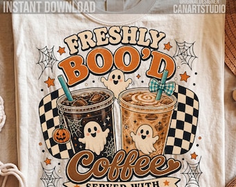 May include: A cream-colored t-shirt featuring a graphic of two iced coffee cups with ghosts, a pumpkin charm, and a checkered pattern. The text reads: FRESHLY BOO'D Coffee SERVED WITH SCREAM & SUGAR. The design includes spiderwebs and orange stars.
