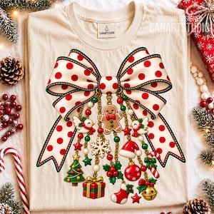 Peut inclure: Un t-shirt couleur crème arborant un grand motif de nœud blanc à pois rouges. Suspendus au nœud, des chaînes dorées sont ornées de breloques de Noël, dont un bonhomme en pain d'épices, un cadeau, un sapin, une chaussette et une boule rouge.