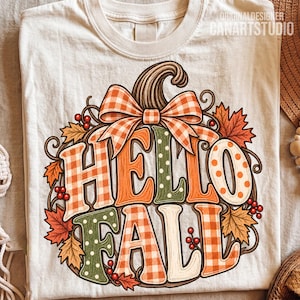 May include: A cream-colored t-shirt featuring a graphic of a pumpkin with a plaid bow. The text HELLO FALL is written in large, multi-patterned letters. The design is surrounded by autumn leaves, swirling vines, and small red berries.