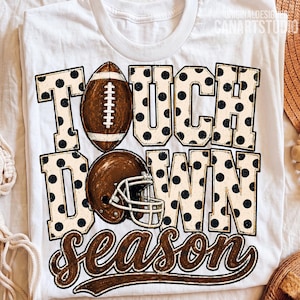 May include: A white t-shirt featuring the text TOUCH DOWN SEASON in large block letters with a black polka dot pattern. A brown football replaces the letter O in TOUCH, and a brown football helmet is positioned over the letter O in DOWN.
