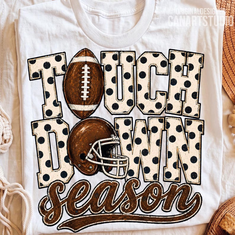May include: A white t-shirt featuring the text TOUCH DOWN SEASON in large block letters with a black polka dot pattern. A brown football replaces the letter O in TOUCH, and a brown football helmet is positioned over the letter O in DOWN.