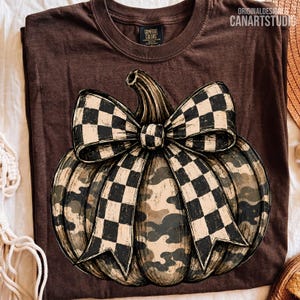 May include: A dark brown t-shirt featuring a large graphic of a pumpkin with a camouflage pattern and a prominent black and white checkered bow. The shirt has a Comfort Colors brand label at the neck and the text ORIGINALDESIGN CANARTSTUDIO.