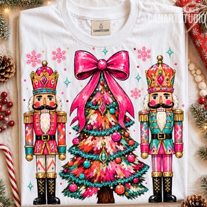 May include: A white t-shirt featuring a graphic print of two nutcrackers flanking a central Christmas tree topped with a large pink bow. The design includes pink and teal snowflakes, ornaments, and stars, with a Canartstudio label at the neck.