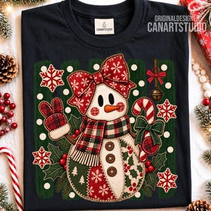 Puede incluir: Una camiseta azul marino oscuro con el gráfico de un muñeco de nieve que lleva un lazo y una bufanda de cuadros rojos. El muñeco sostiene un bastón de caramelo y lleva manoplas de cuadros. El diseño incluye copos de nieve blancos, hojas de acebo y tres botones marrones en el cuerpo. El texto CANARTSTUDIO aparece en la parte superior.