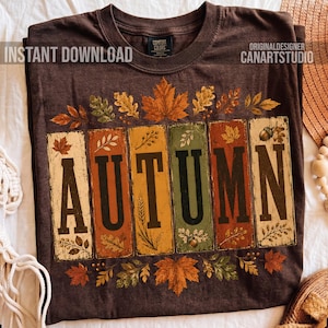 May include: A dark brown t-shirt featuring the word AUTUMN in large black letters. Each letter is set against a vertical rectangular panel in shades of cream, orange, yellow, and green. The design is decorated with various autumn leaves and acorns.