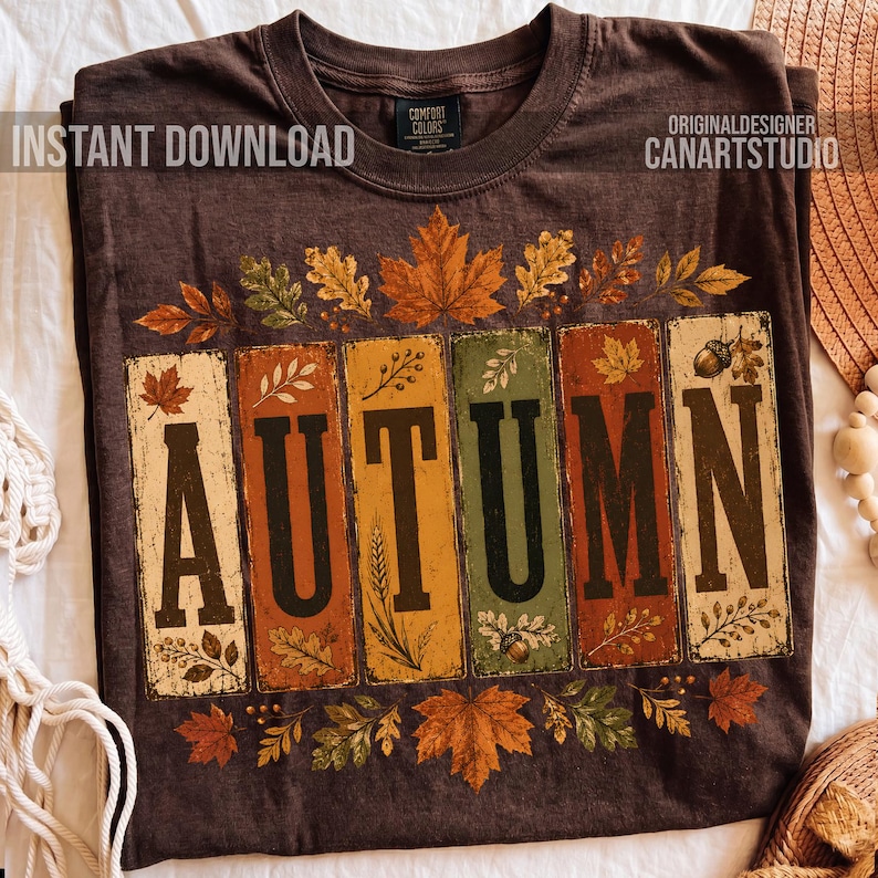 May include: A dark brown t-shirt featuring the word AUTUMN in large black letters. Each letter is set against a vertical rectangular panel in shades of cream, orange, yellow, and green. The design is decorated with various autumn leaves and acorns.