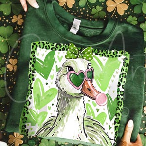May include: Green sweatshirt featuring a graphic of a duck wearing heart-shaped sunglasses and a bow. The duck is surrounded by green hearts and a leopard print border. The sweatshirt is folded.