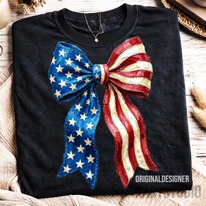 May include: Black t-shirt with a large bow design in patriotic colours. The bow features blue and white stars on one side and red and white stripes on the other. The text "ORIGINALDESIGNER" is at the bottom.