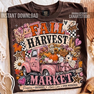 May include: A brown t-shirt featuring a graphic of a pink vintage truck filled with pumpkins, sunflowers, and flowers. The text reads FALL HARVEST MARKET, PUMPKINS, HAYRIDES, APPLE CIDER, and OPEN WEEKENDS, decorated with bows, stars, and hearts.