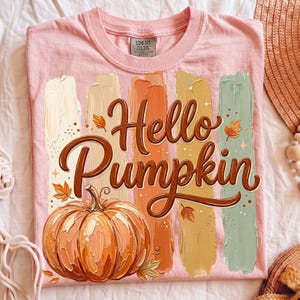 Puede incluir: Camiseta rosa claro con la frase "Hello Pumpkin" en marr&oacute;n y una ilustraci&oacute;n de calabaza. El dise&ntilde;o presenta pinceladas verticales en colores oto&ntilde;ales. Marca Comfort Colors.