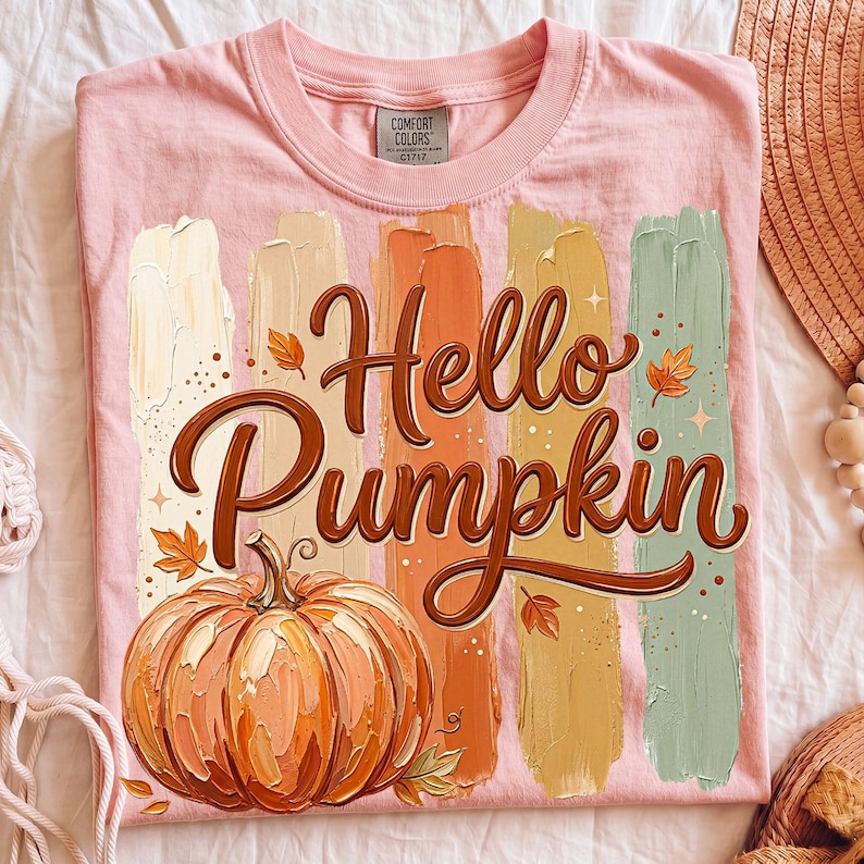 Puede incluir: Camiseta rosa claro con la frase "Hello Pumpkin" en marr&oacute;n y una ilustraci&oacute;n de calabaza. El dise&ntilde;o presenta pinceladas verticales en colores oto&ntilde;ales. Marca Comfort Colors.