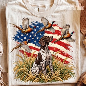 May include: A cream-colored t-shirt featuring a graphic print of a brown and white spotted dog sitting in a field of wheat. Three pheasants are flying above the dog, set against a backdrop of a stylized American flag with red, white, and blue.