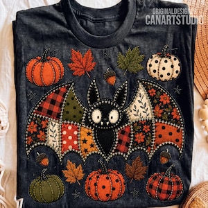 Puede incluir: Una camiseta gris oscuro con un gráfico central de un murciélago con alas estilo patchwork en varios patrones otoñales. El diseño incluye calabazas naranjas, verdes y de lunares, hojas de arce y bellotas. El texto ORIGINALDESIGN CANARTSTUDIO es visible.