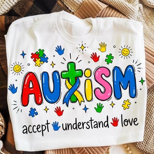 以下が含まれることがあります： カラフルな文字で「AUTISM」と書かれた白いTシャツ。パズルのピース、手形、星、そして「accept understand love」という言葉がデザインされています。クルーネックのシャツです。