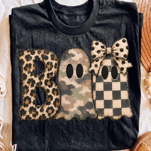 May include: A black short-sleeve t-shirt featuring the word BOO in large letters. The letter B has a leopard print, the middle ghost has a camouflage pattern, and the final ghost has a black and white checkerboard pattern with a polka dot bow.