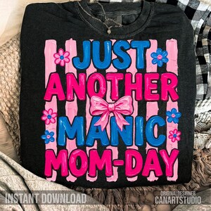 Op de afbeelding: Donkergrijze sweatshirt met de tekst "JUST ANOTHER MANIC MOM-DAY" in roze en blauw, met een roze strik en bloemaccenten. De woorden zijn geplaatst tegen roze verticale strepen. De zin "INSTANT DOWNLOAD" staat onderaan.