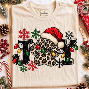May include: A beige t-shirt featuring the word JOY in black, with the letter O replaced by a leopard-print paw print wearing a red Santa hat. A string of colorful Christmas lights wraps around the letters, accented by red and green snowflakes.