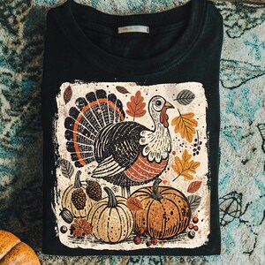 Pode incluir: Camisola preta de gola redonda com um design de A&ccedil;&atilde;o de Gra&ccedil;as. O design inclui um peru, ab&oacute;boras e folhas de outono em tons de laranja, castanho e bege. A camisola est&aacute; dobrada sobre uma superf&iacute;cie estampada.