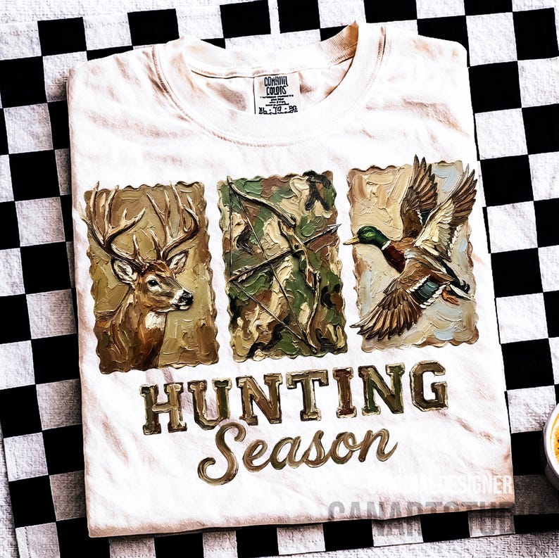 May include: Cream-colored t-shirt with a hunting theme. The shirt features three framed images: a deer, a bow and arrow, and a duck in flight. The words "HUNTING SEASON" are printed in a gold-colored, stylized font.