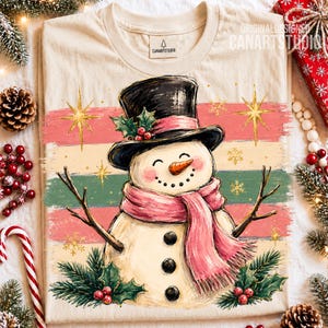 Może przedstawiać: Kremowy t-shirt z grafiką bałwana w czarnym cylindrze i r&oacute;żowym szaliku. Bałwan ma marchewkowy nos, gałązkowe ramiona i trzy czarne guziki, a całość ozdobiona jest liśćmi ostrokrzewu, jagodami oraz złotymi gwiazdkami na tle kolorowych pas&oacute;w.