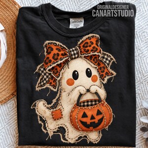 May include: A black t-shirt featuring a cream-colored ghost graphic with a frayed edge applique. The ghost wears a large leopard print and black-and-white checkered bow, and holds an orange jack-o'-lantern bucket. Text reads CANARTSTUDIO.