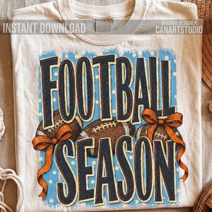 K&ouml;nnte beinhalten: Ein cremefarbenes T-Shirt mit einem Aufdruck, der den Text FOOTBALL SEASON in dunkelblauen Buchstaben zeigt. Das Design umfasst drei braune Footballs, die jeweils mit einer orangefarbenen Schleife verziert sind, auf hellblauem Grund.
