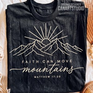 May include: A charcoal gray t-shirt featuring a white line art graphic of a sun rising over mountain peaks. The text reads "FAITH CAN MOVE mountains" and "MATTHEW 17:20" in a mix of block and script fonts. The top includes "ORIGINALDESIGNER CANARTSTUDIO".