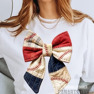 Puede incluir: Un gran lazo decorativo con un diseño de patchwork que incorpora elementos de béisbol. El lazo incorpora telas rojas, crema y azul marino, con costuras rojas que imitan las costuras de béisbol. El lazo está sujeto a una camiseta blanca.