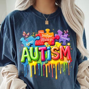 May include: A dark blue t-shirt with a colorful graphic design of the word "AUTISM" with puzzle pieces and butterflies. The puzzle pieces have text that reads "Unique", "Intelligent & Always", and "Totally Sometimes Mysterious".