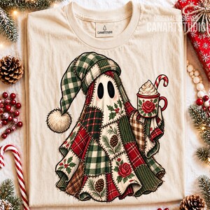 May include: A cream-colored t-shirt featuring a graphic of a ghost wearing a patchwork quilt-style hat and cloak. The ghost holds a mug with a red rose design, whipped cream, and a candy cane. The quilt pattern includes red, green, and brown.