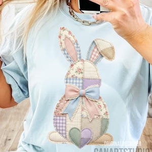 May include: Light blue t-shirt featuring a patchwork bunny design. The bunny has floral, gingham, and solid fabric patches in pink, blue, purple, and beige. A pink and blue bow adorns the bunny's neck, and a heart is at the bottom. Text on the bottom right reads "CANARTSTUDIO".