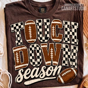 May include: A dark brown t-shirt featuring the text TOUCH DOWN season in a mix of black and white checkered patterns and brown football-themed graphics. The design includes a brown football illustration at the bottom right corner.