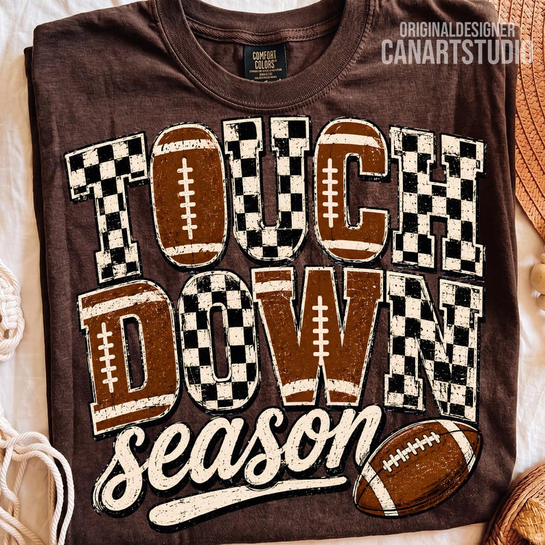May include: A dark brown t-shirt featuring the text TOUCH DOWN season in a mix of black and white checkered patterns and brown football-themed graphics. The design includes a brown football illustration at the bottom right corner.