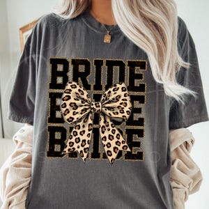 May include: Grey t-shirt with the text "Bride Tribe" in black with a gold glitter outline. A large leopard print bow is centred on the shirt.