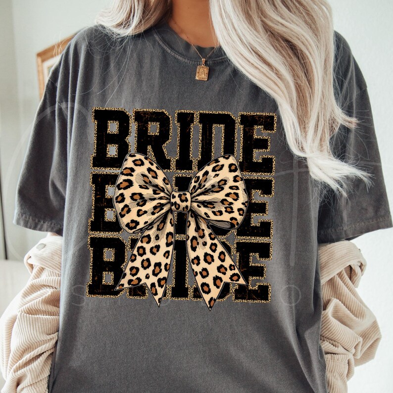 May include: Grey t-shirt with the text "Bride Tribe" in black with a gold glitter outline. A large leopard print bow is centred on the shirt.