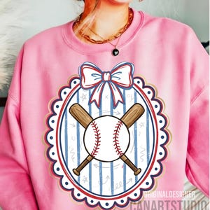 May include: Pink sweatshirt featuring a baseball design. The design includes a baseball with red stitching, crossed wooden bats, and a bow. The design is set within a scalloped oval frame with blue and gold accents. The text "CANARTSTUDIO" is visible.