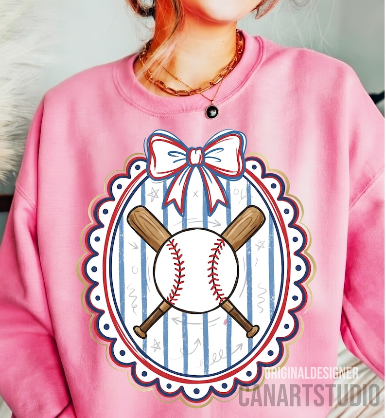 May include: Pink sweatshirt featuring a baseball design. The design includes a baseball with red stitching, crossed wooden bats, and a bow. The design is set within a scalloped oval frame with blue and gold accents. The text "CANARTSTUDIO" is visible.