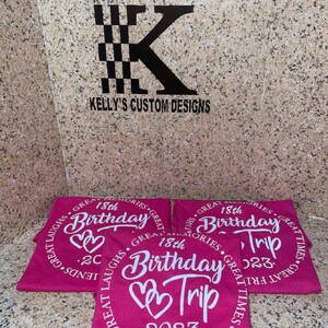 May include: Three pink t-shirts with white text that says "Great Memories, Great Laughs, Great Times" and "18th Birthday Trip 2023" with two hearts.