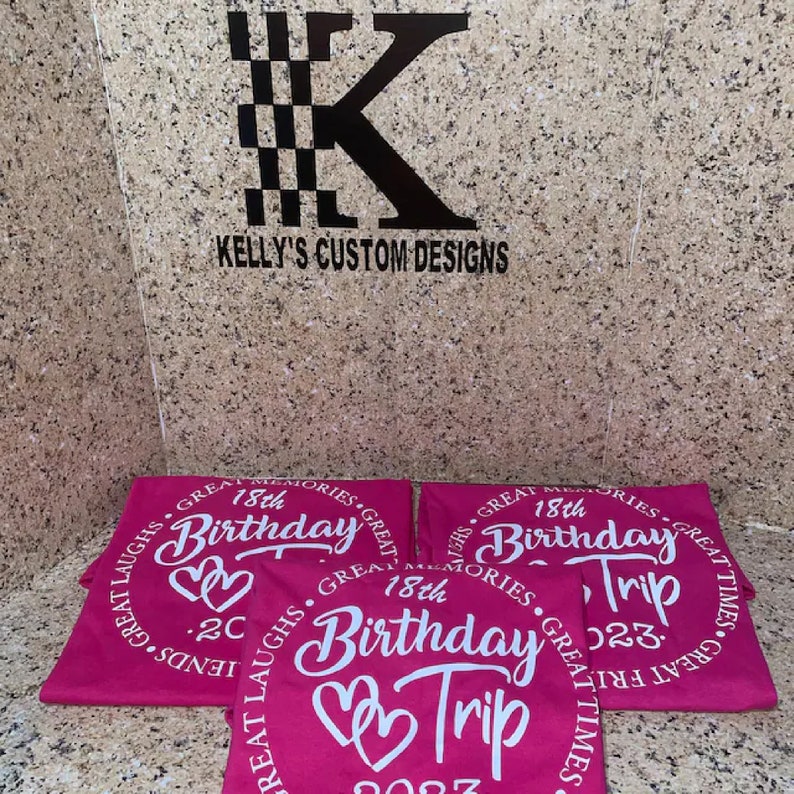 May include: Three pink t-shirts with white text that says "Great Memories, Great Laughs, Great Times" and "18th Birthday Trip 2023" with two hearts.